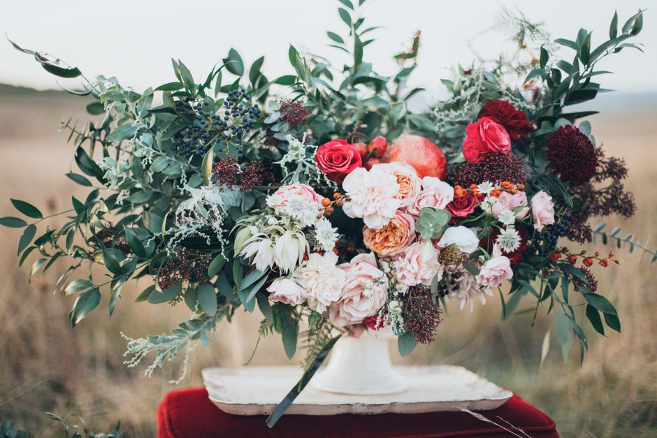A large floral arrangement featuring a variety of fresh flowers, including blush pink and cream roses, deep red dahlias, white carnations, and small pink blooms, interwoven with lush greenery such as