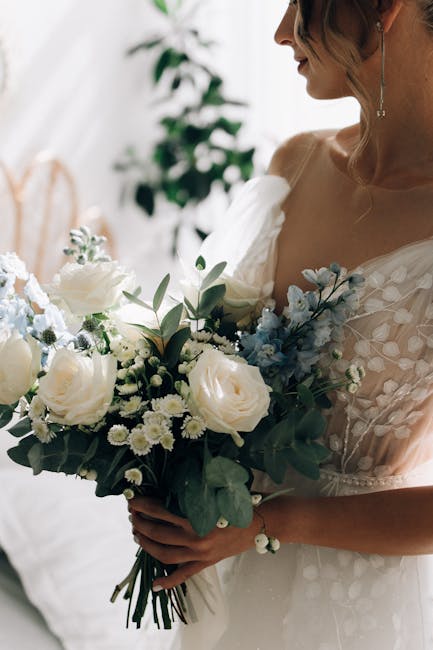 A woman in a white dress holding a bouquet of fresh flowers, featuring white roses, small white daisies, light blue accent flowers, and green foliage. The bouquet is arranged with a mix of lush blooms