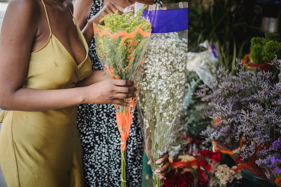 Close-up of a person in a yellow satin slip dress holding a tall bouquet of delicate, small green and white flowers wrapped in clear plastic with orange accents, positioned in front of a vibrant flora