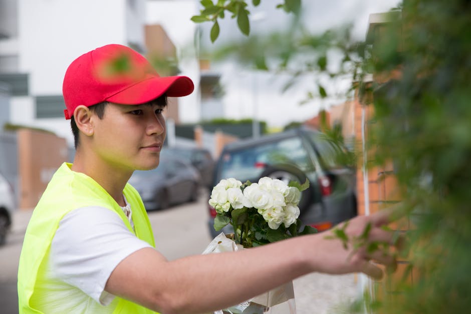 A delivery person wearing a red cap and a fluorescent yellow vest is placing a bouquet of fresh white hydrangeas against a green leafy shrub. The bouquet is arranged in a simple cluster, showcasing vi