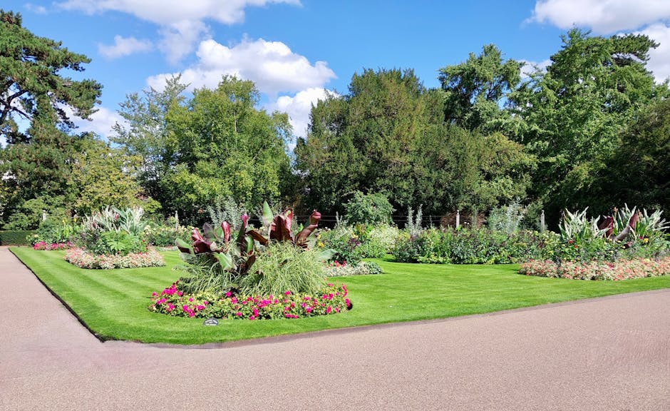 A scenic park scene featuring neatly maintained flower beds with a variety of colorful blooms, including pink, purple, and white flowers, arranged around lush green shrubbery and large flowering plant