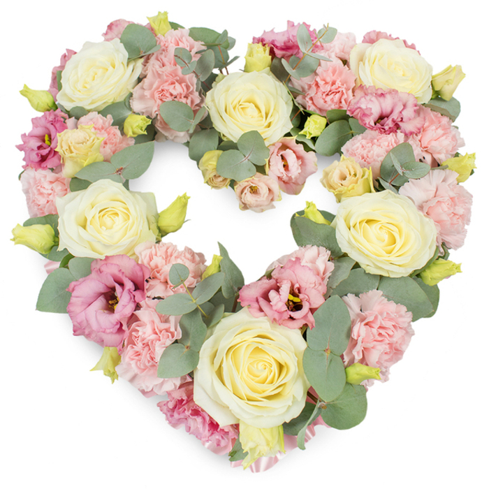 Displayed on a clear white background, this heart-shaped funeral wreath forms a complete, symmetrical outline of love crafted entirely from pastel flowers and cool greenery. The shape is unmistakably a heart, yet softened by the abundance of blooms that spill into one another, creating a plush, almost cushion-like surface. Several large roses in shades of cream and very pale yellow are placed with intention around the wreath, their tightly layered, velvety petals unfolding in elegant spirals that signal purity, devotion, and enduring affection. Filling the spaces between these focal blooms are clusters of light pink carnations, each small flower made up of many frilled petals that lend a gentle, textured contrast and an airy feel. Deeper pink to mauve lisianthus blooms are sprinkled thoughtfully throughout, some fully open, others just beginning to unfurl, adding both tonal depth and a graceful, romantic character to the tribute. Tiny yellow-green buds nestle between the petals, suggesting youth, renewal, and the ongoing cycle of life. Silvery-green eucalyptus leaves weave across the design, their rounded, matte foliage resting lightly against the softer pastels, giving the wreath a subtle, contemporary freshness. The flowers sit densely together, so the heart feels rich, full, and carefully composed, much like the bespoke tributes crafted by experienced florists serving families in Grove Park. This arrangement would be fitting for a farewell service, a memorial gathering, or a celebration of life, offering a gentle focal point that speaks of remembrance, gratitude, and the quiet strength of love.